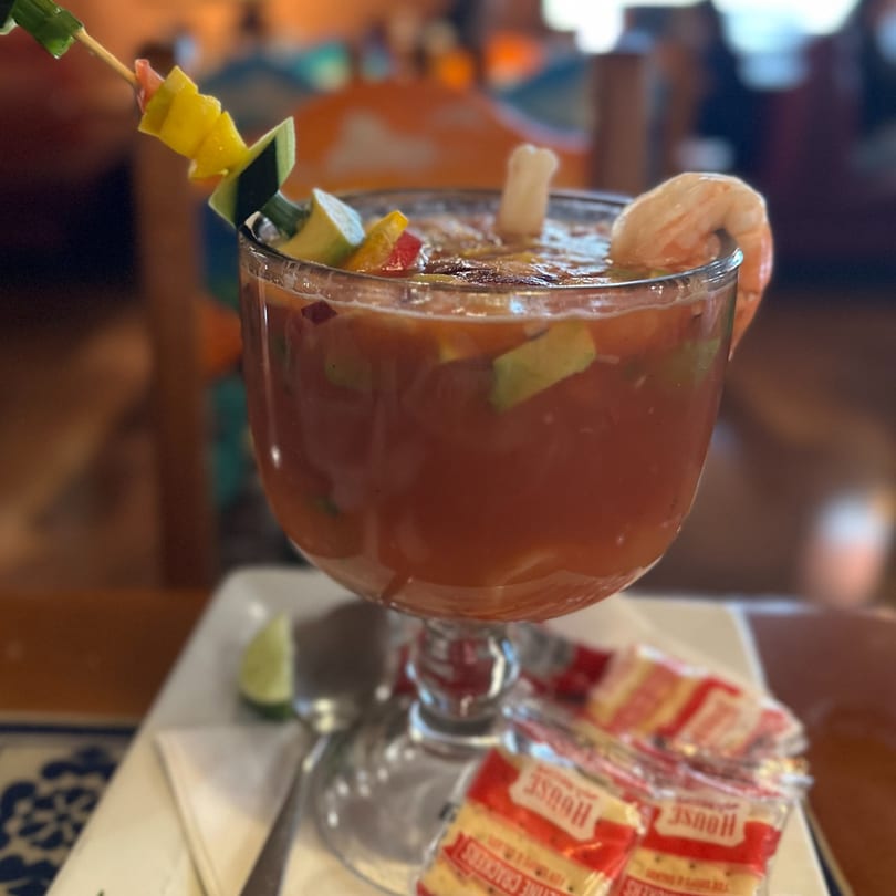 A large glass of Michelada, garnished with shrimp, lime, and a vegetable skewer, with crackers.