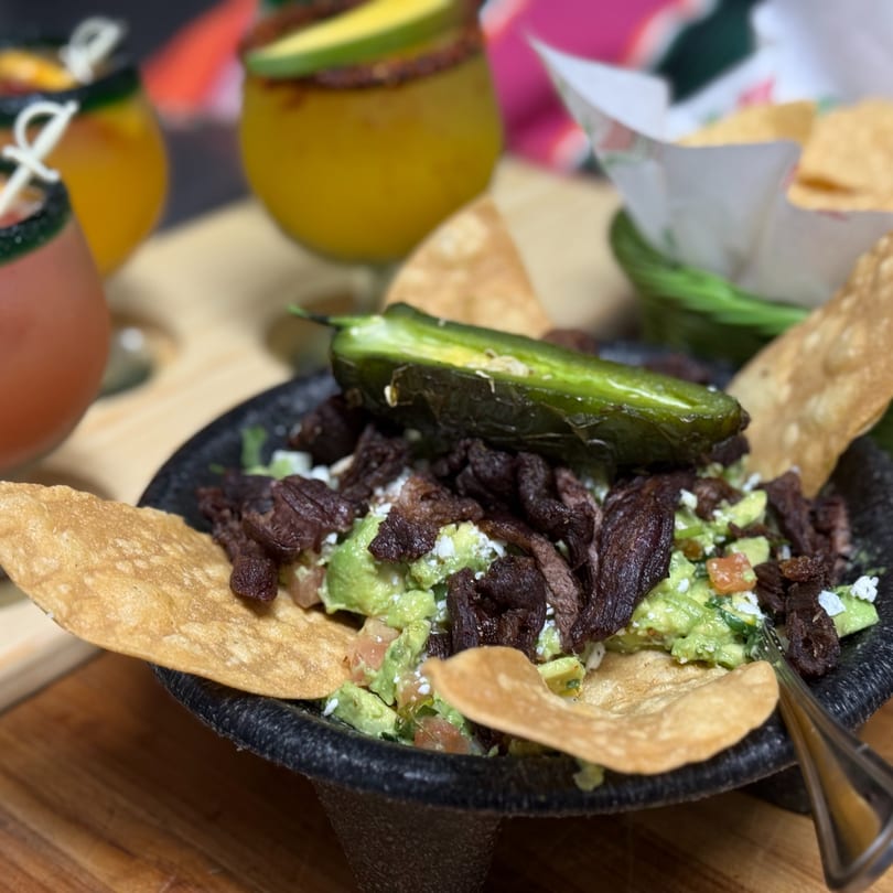 Guacamole with steak, a grilled jalapeño, and tortilla chips in a dark bowl, with drinks in the background.