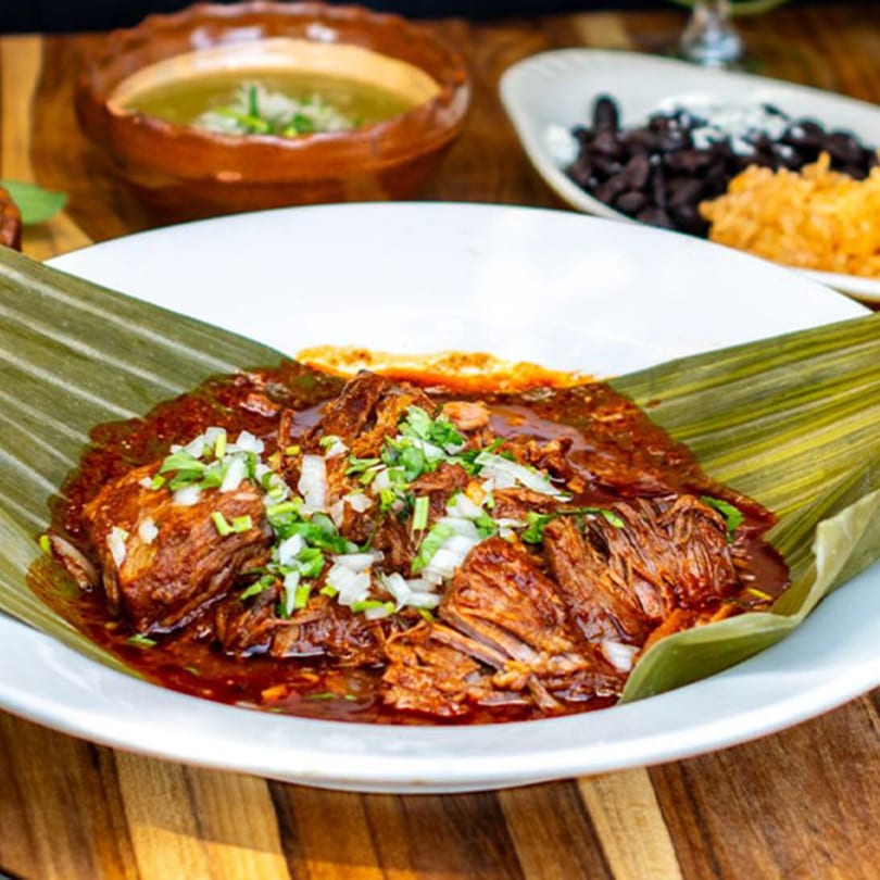 Shredded meat in red sauce with onions and cilantro, served on a banana leaf with sides.