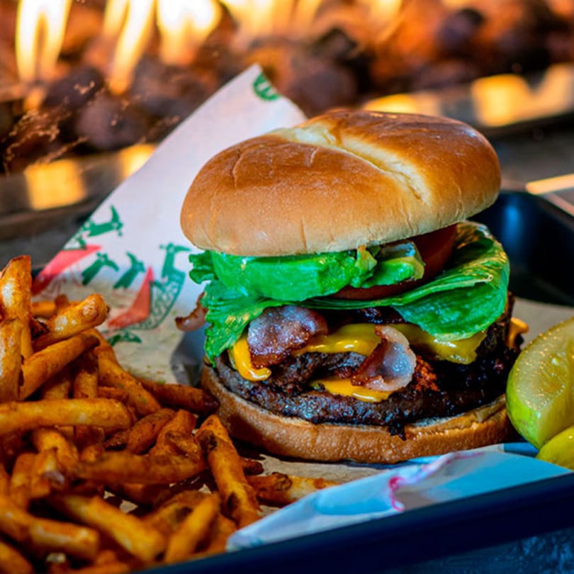 A bacon cheeseburger with fries and pickles on a tray, with a fire pit in the background.