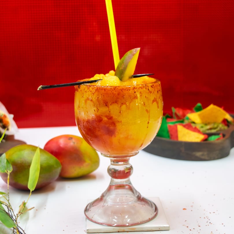 A vibrant mango drink in a goblet, garnished with mango slices and a yellow straw, against a red background.
