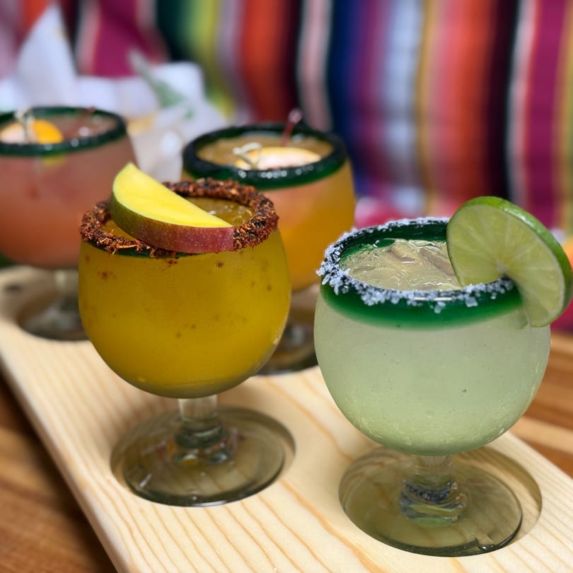 A flight of four colorful margaritas, including mango and lime, on a wooden tray.