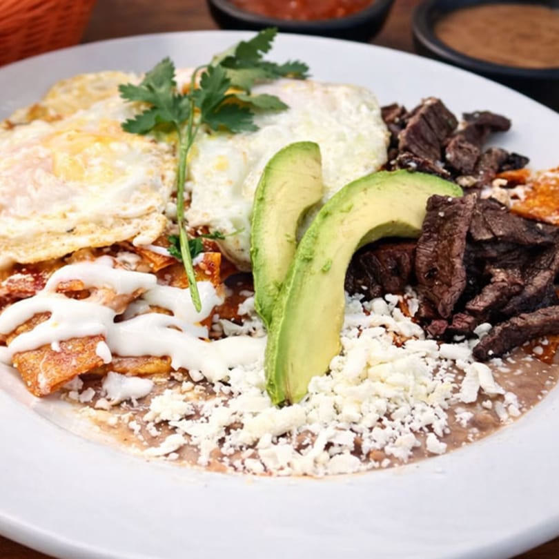 Chilaquiles with fried eggs, steak, avocado, crumbled cheese, and crema on a white plate.