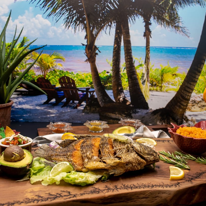 Whole fried fish with avocado, salsa, and rice on a wooden board, with a beach backdrop.