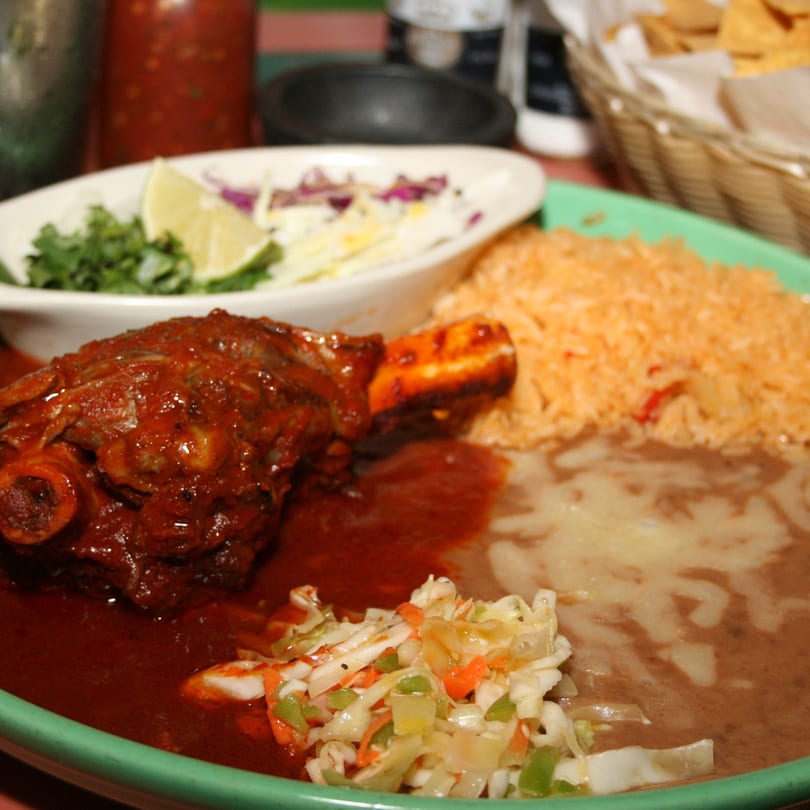 Braised lamb shank with red sauce, rice, refried beans, and coleslaw on a green plate.