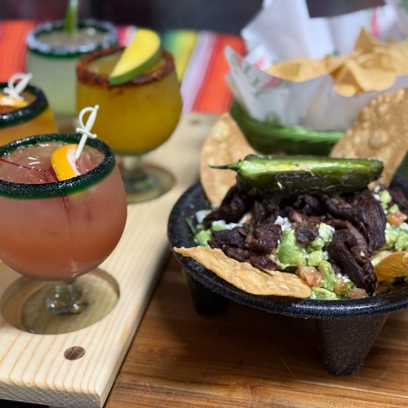 A molcajete bowl with steak, guacamole, and a jalapeño, next to a flight of margaritas.