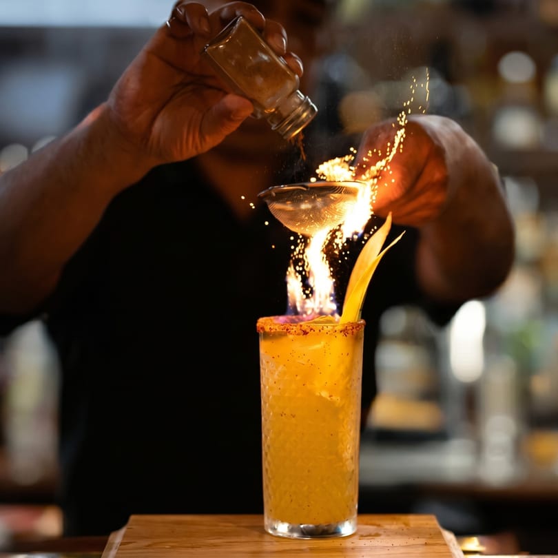 Bartender igniting spices over a tall, garnished cocktail, creating a fiery display.