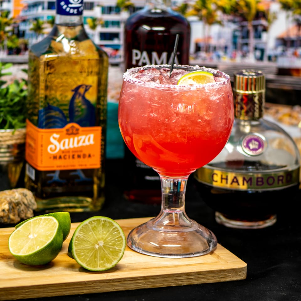 A vibrant red margarita with a salted rim and lime wedge, surrounded by liquor bottles and fresh limes.
