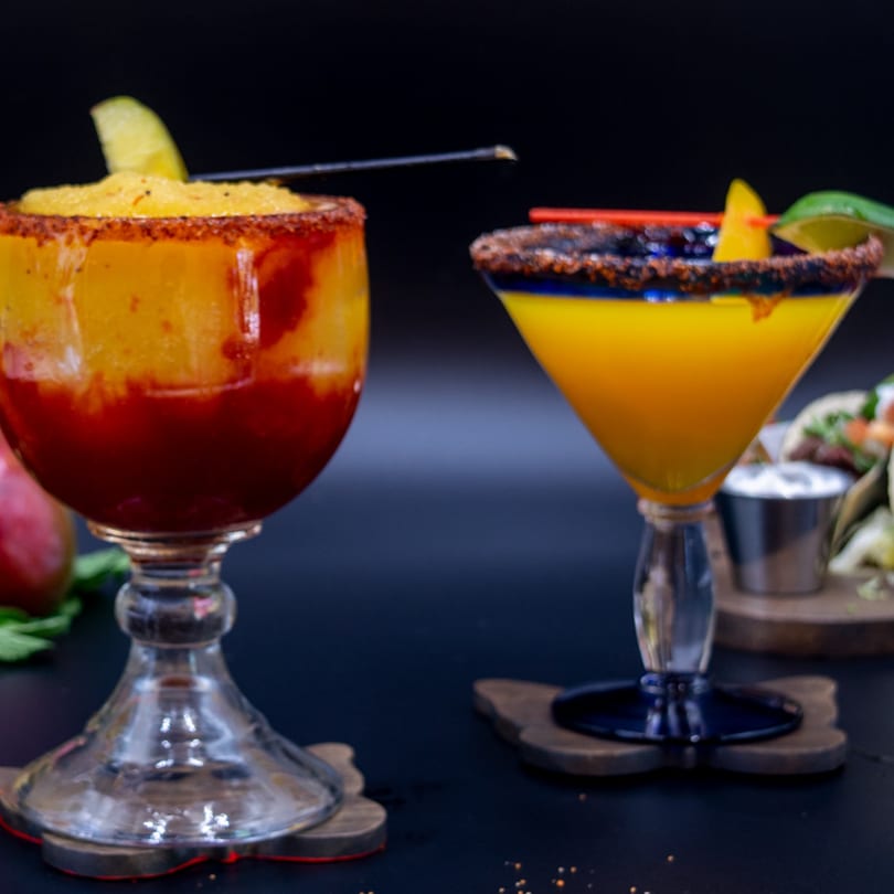 Two vibrant mango drinks with chili-lime rims and garnishes, with tacos blurred in the background.