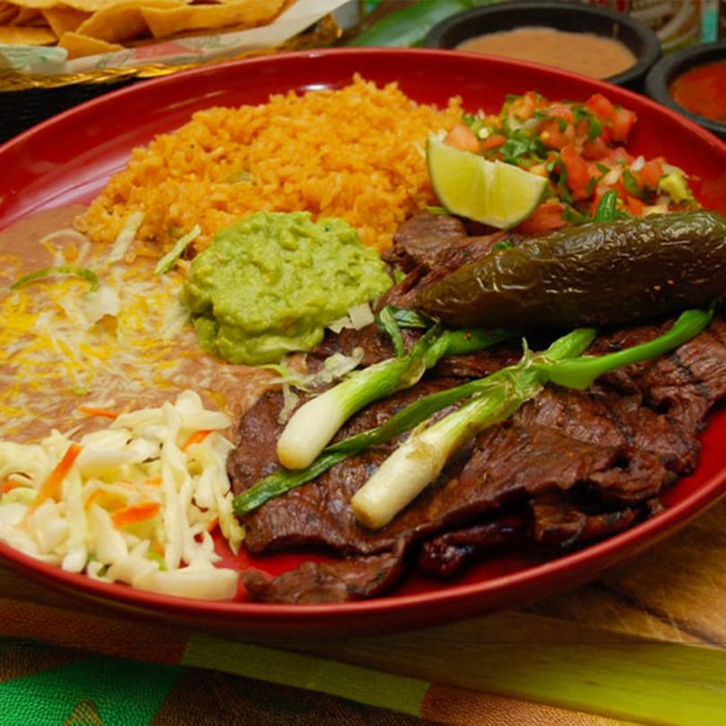 A red plate of carne asada with rice, beans, guacamole, pico de gallo, and grilled scallions.