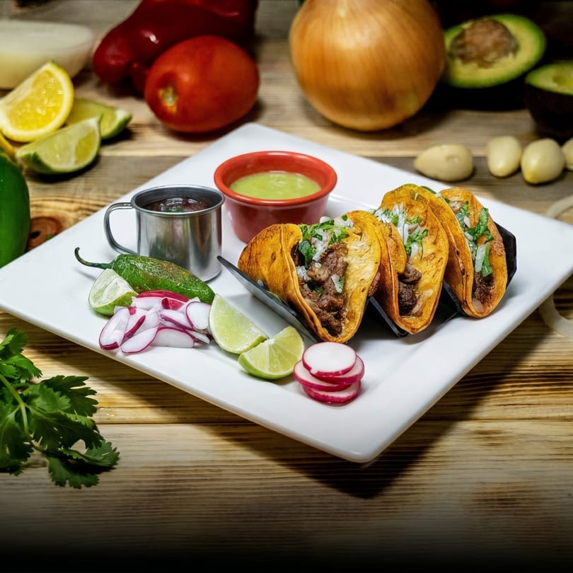 Three birria tacos with lime, radish, jalapeño, and dipping sauces on a white plate.