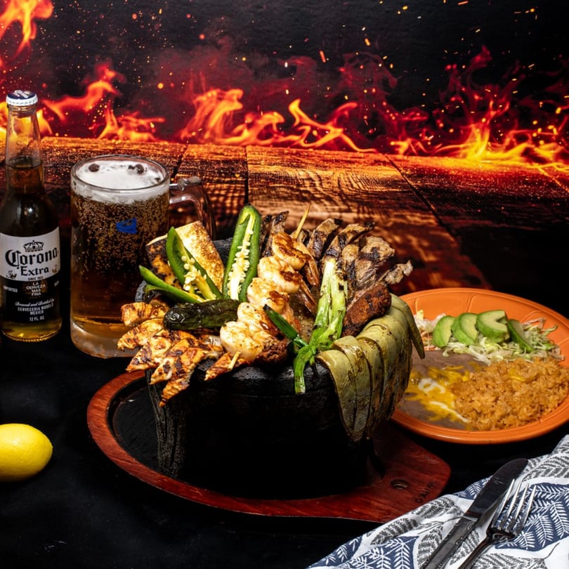 A sizzling molcajete with grilled meats, shrimp, and vegetables, served with rice, beans, and beer.