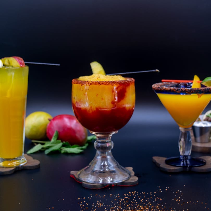 Three colorful mango drinks, two with chili rims, on a dark surface with fresh mangoes and mint.