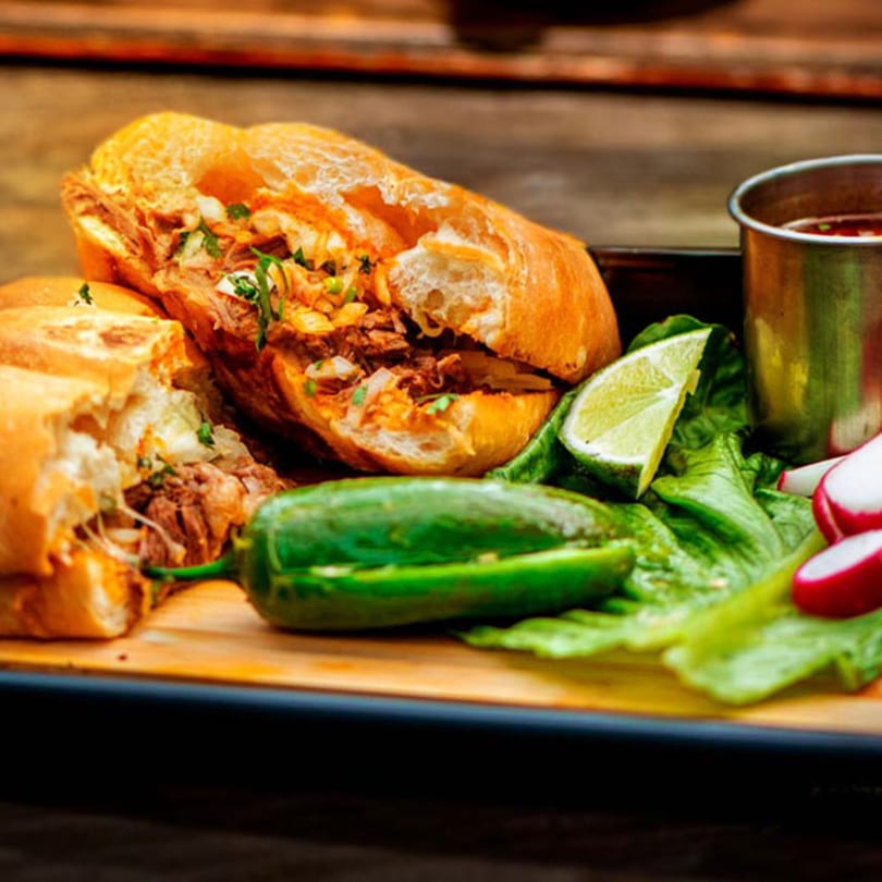 Two halves of a Mexican torta sandwich with meat, cilantro, and onions, served with lime, jalapeño, radishes, and a cup of consommé.