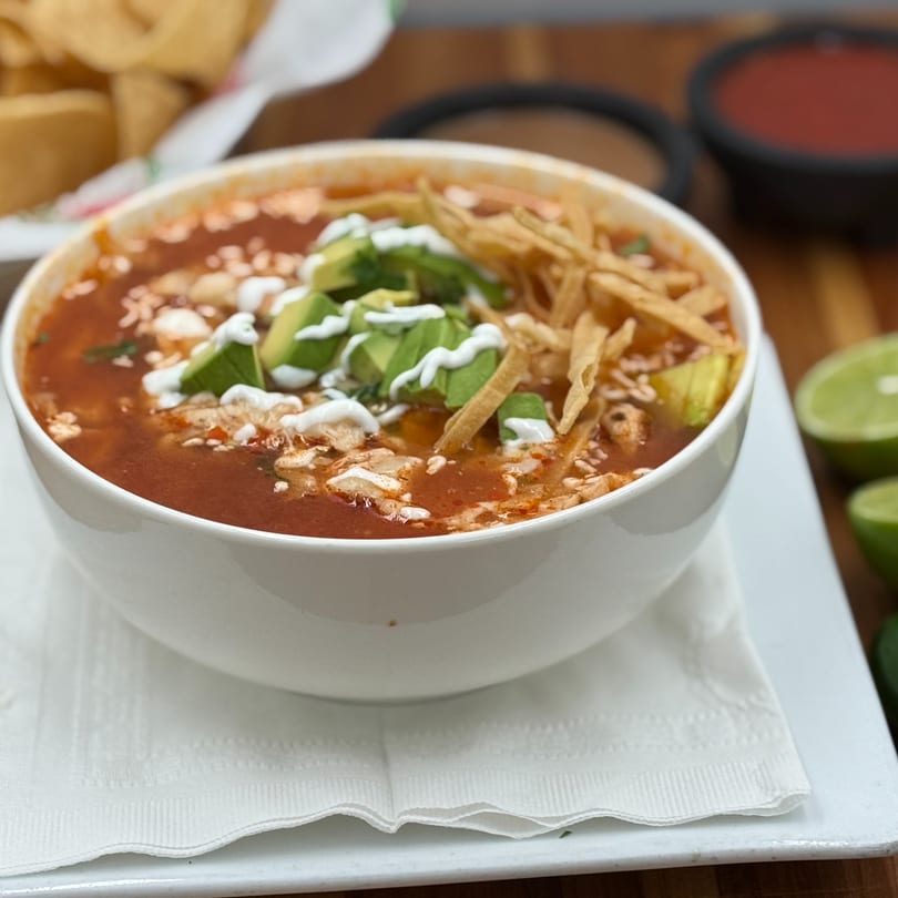 A white bowl of red soup with avocado, sour cream, and tortilla strips on a white plate.