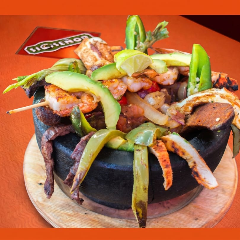 A large molcajete filled with grilled meats, shrimp, avocado, onions, and peppers, on a wooden board.
