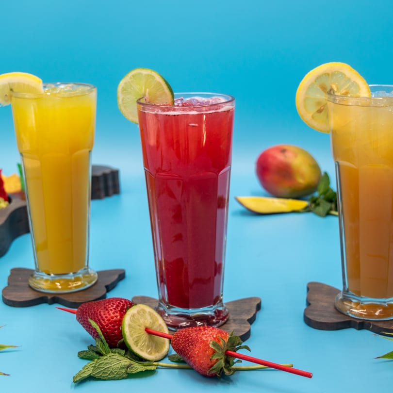 Three colorful fruit drinks with garnishes, including strawberries, lime, and mango, on a blue background.
