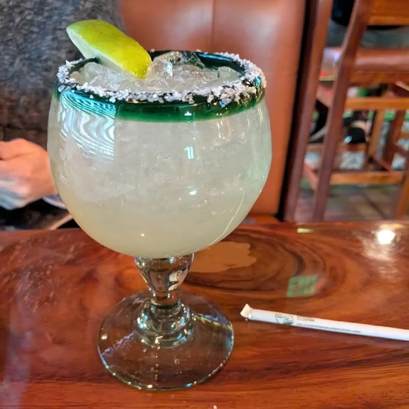 A large margarita with a salted rim and lime wedge sits on a wooden table.