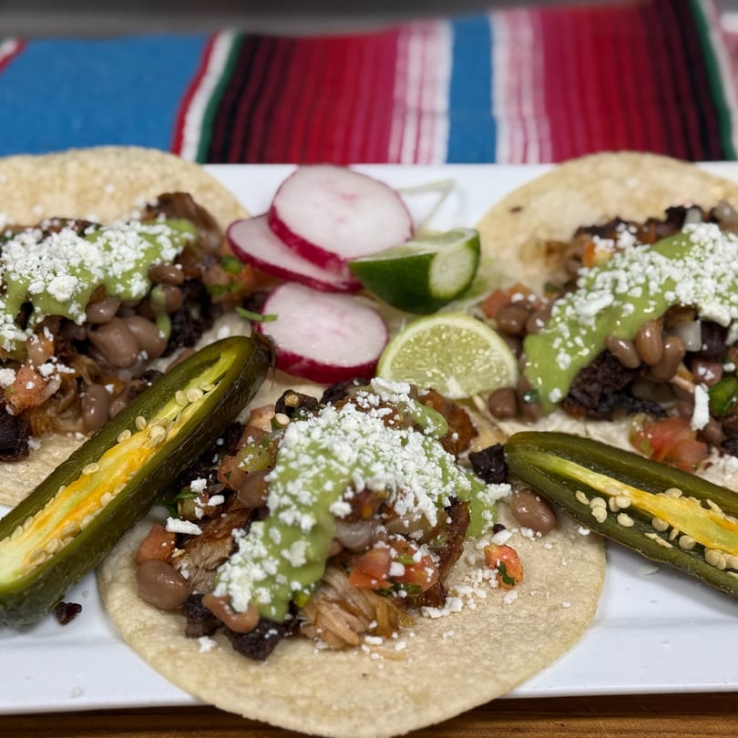 Three tacos on a white plate, topped with meat, beans, avocado sauce, cheese, radishes, and jalapeños.