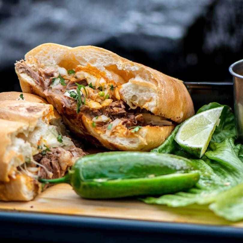 Two halves of a meat-filled sandwich with cilantro, onions, a jalapeño, and lime on lettuce.